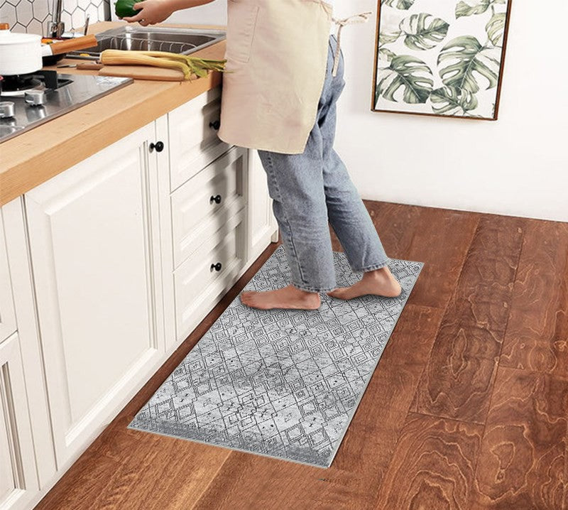 Alfombrilla 'Anissa' en una cocina, con el pie de una persona sobre ella. El diseño geométrico en tonos grises y negros con patrones de rombos y detalles intrincados complementa el ambiente de la cocina. La alfombra es lavable en lavadora y tiene una base antideslizante.