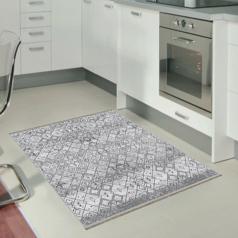 Pequeña alfombra 'Anissa' en una cocina. El diseño geométrico en tonos grises y negros con patrones de rombos y detalles intrincados complementa el ambiente de la cocina. La alfombra es lavable en lavadora y tiene una base antideslizante.