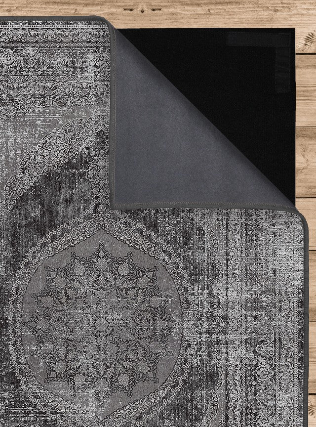 Vista de la alfombra 'Aria' mostrando el frente y el reverso de la capa superior con su diseño intrincado de medallón y bordes ornamentales en tonos de gris, negro y blanco. También se aprecia la capa inferior antideslizante, que asegura estabilidad. La alfombra es lavable en lavadora y no suelta pelusa.