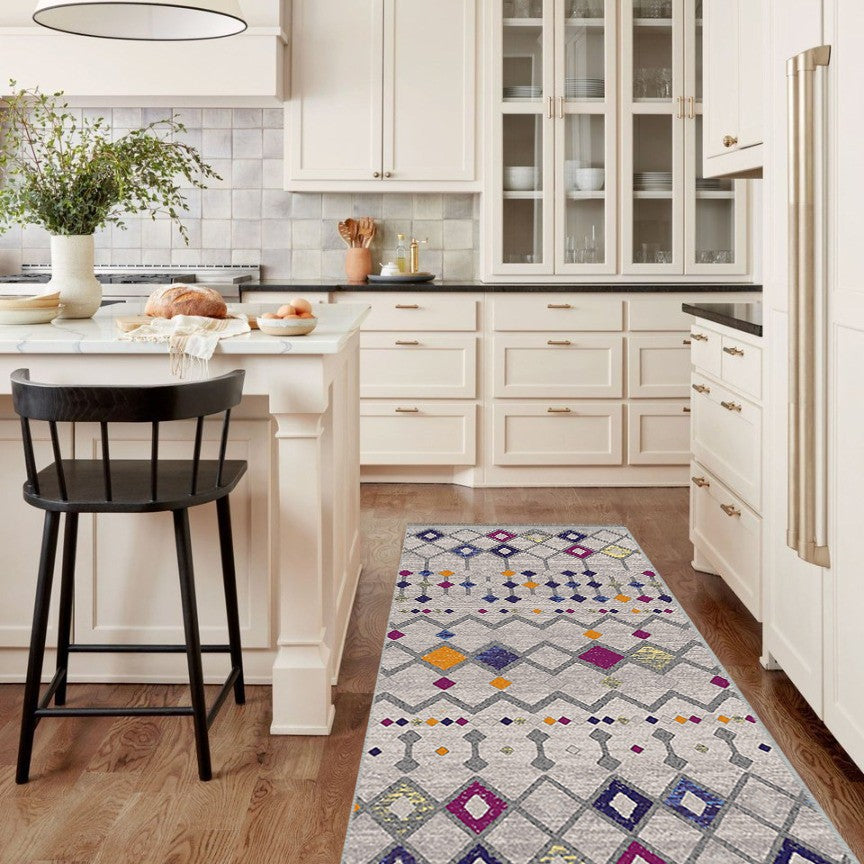 Alfombra 'Nadia' en una cocina, aportando un toque moderno y vibrante con su diseño en tonos púrpura, naranja, gris y blanco. Esta alfombra es lavable en lavadora, facilitando su cuidado en un entorno de alto tráfico.